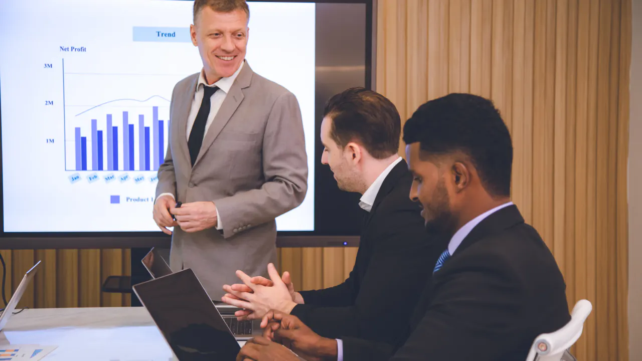 Presenter at a screen with a sales chart addressing two colleagues, illustrating how to influence AE sales