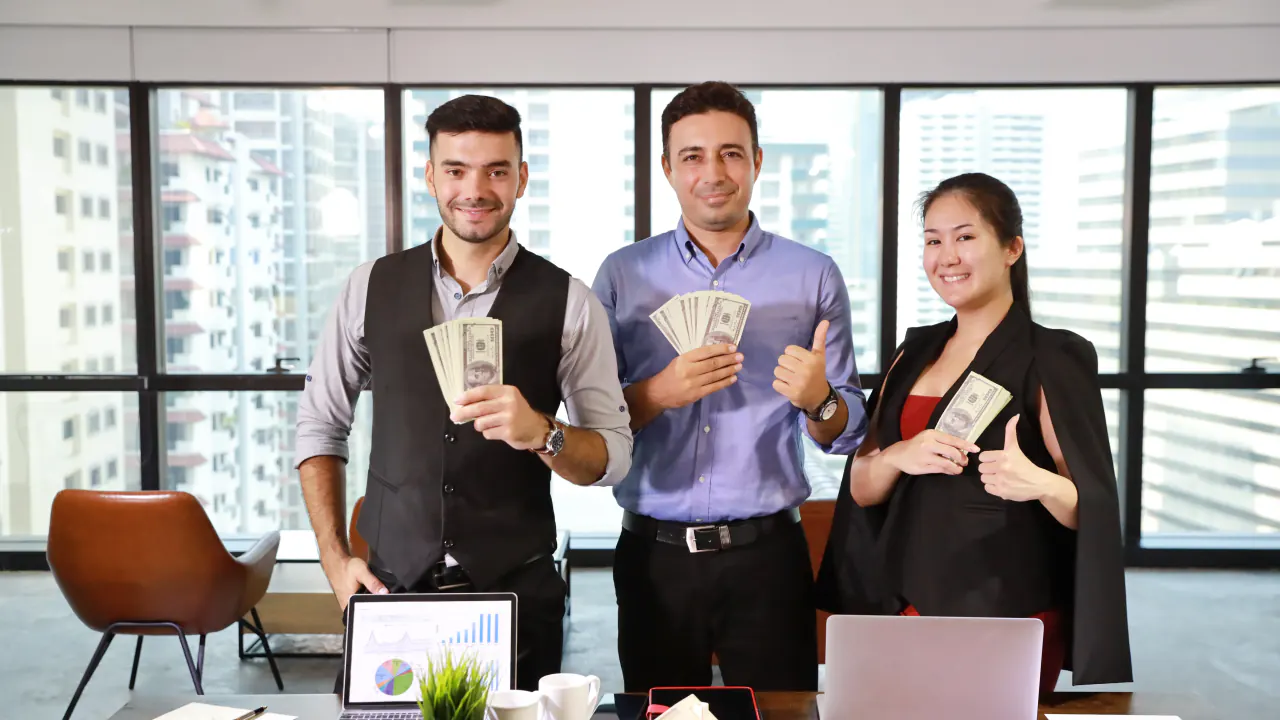 Three colleagues holding cash and giving thumbs up, illustrating on-target earnings (OTE)